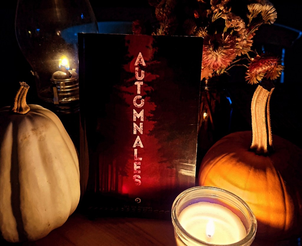 Livre entouré de citrouilles et de chandelles. Sur la couverture, une forêt sombre se découpe sur un fond rouge et des lettre à la verticale forment le mot Automnales.
