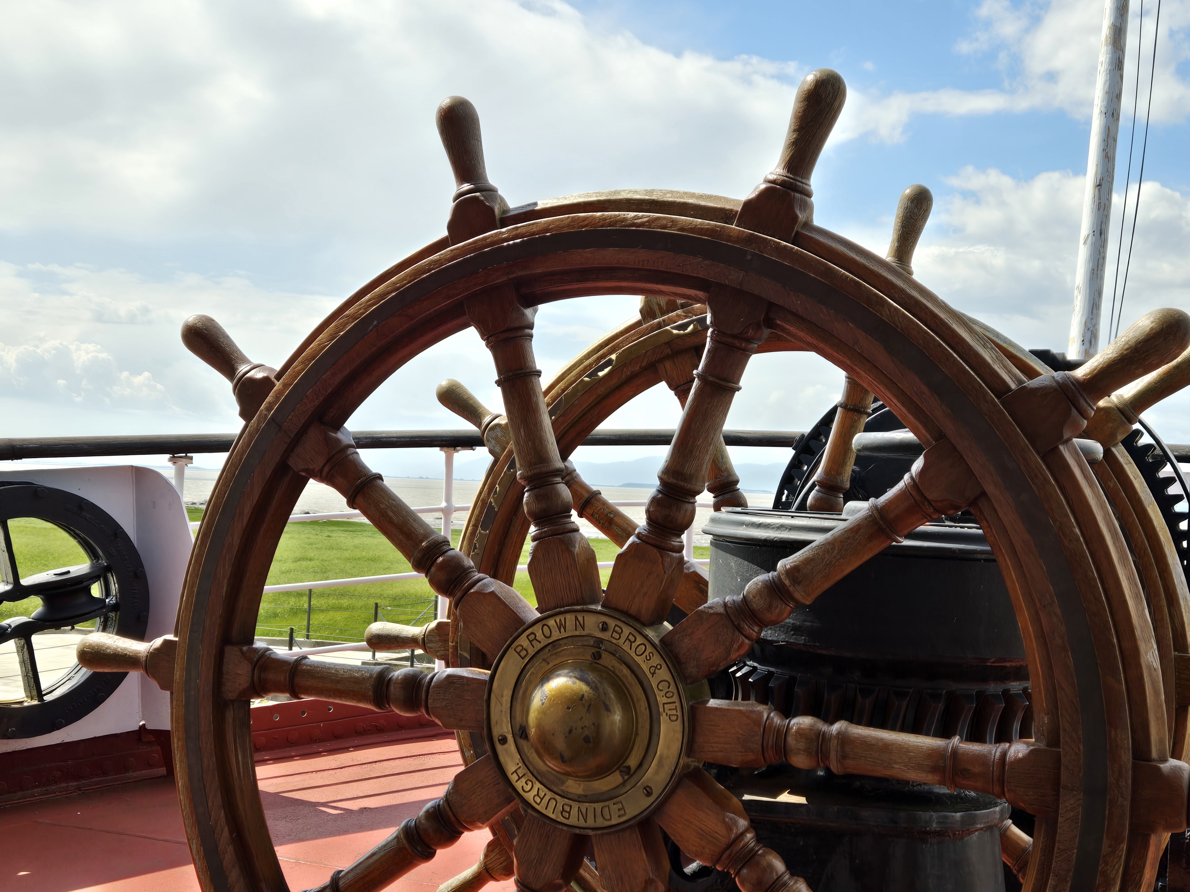 Un ancien gouvernail à double roue en bois avec un centre en laiton sur lequel est écrit Brown Bros & Co Edinburgh. En arrière plan on voit le pont du bateau Ernest Lapointe et le fleuve Saint-Laurent sous un ciel bleu nuageux.