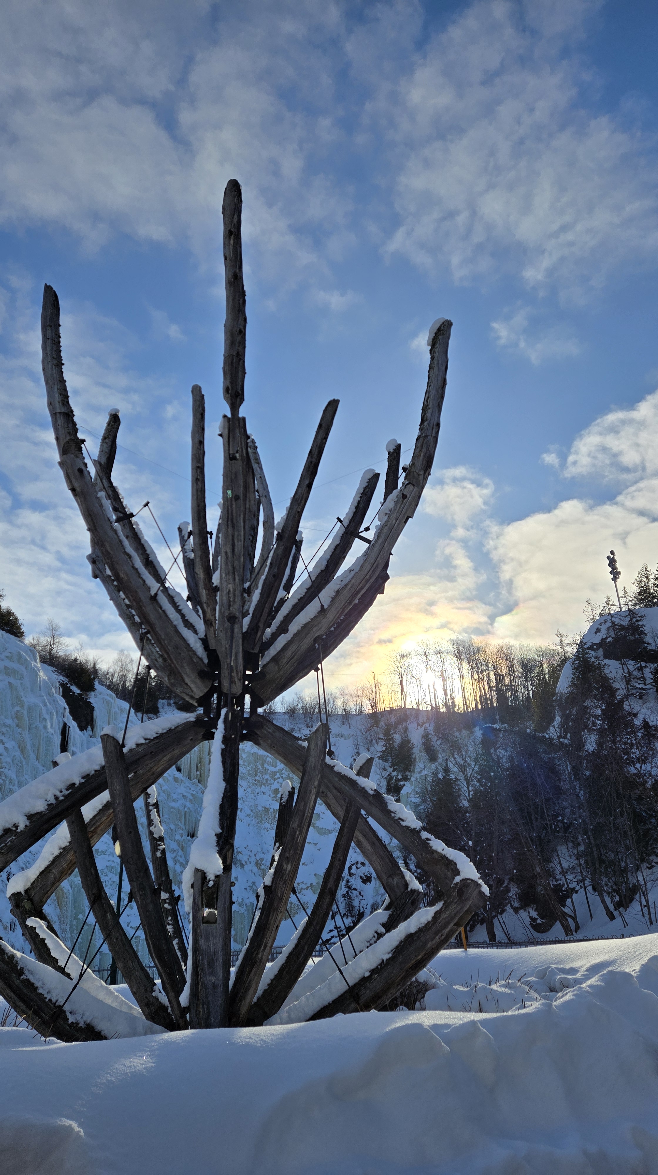 Sculpture faite de morceaux de bois assemblés à la manière d'une main mécanique qui tend ses doigts vers le soleil qui se lève au-dessus de la falaise du parc des chutes