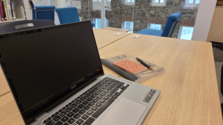 Ordinateur portable et cahier d'écriture sur une table avec en arrière plan une étagère de livres et les murs de pierre de la bibliothèque Françoise-Bédard