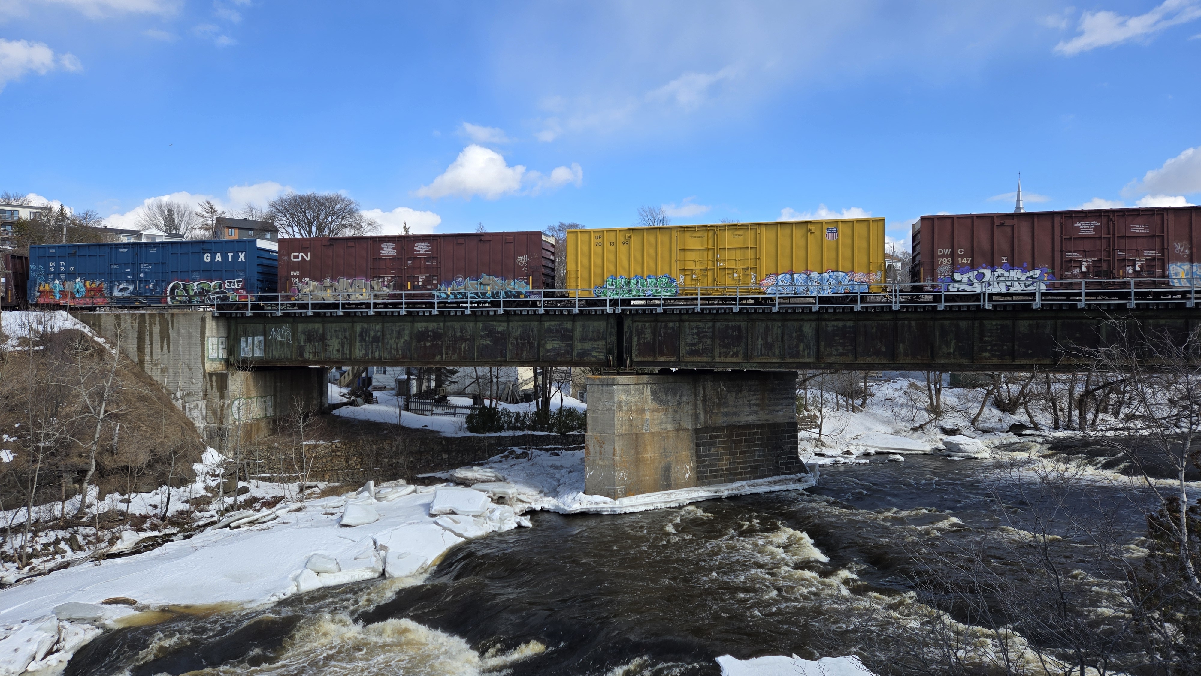 Quatre wagons colorés sur lesquels on a peint des graffitis. Le train passe sur un pont au-dessus des rapides de la rivière du loup partiellement gelée
