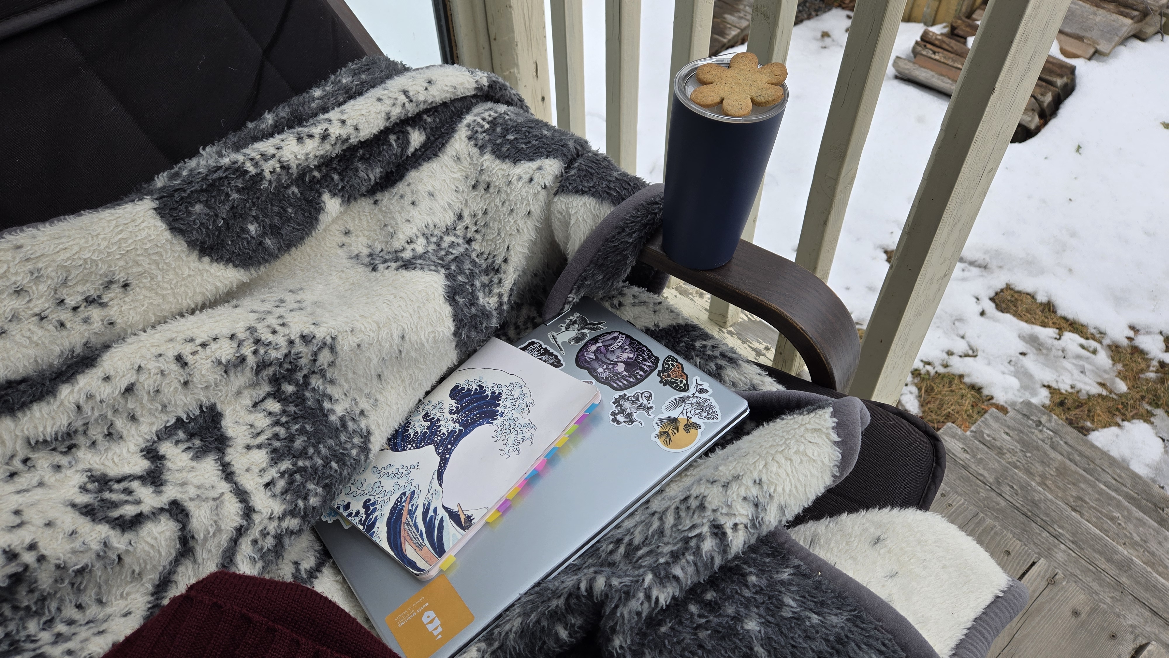 Chaise sur un balcon avec couverture, ordinateur portable, carnet, tasse thermal et biscuit. On voit encore de la neige en bas du balcon.