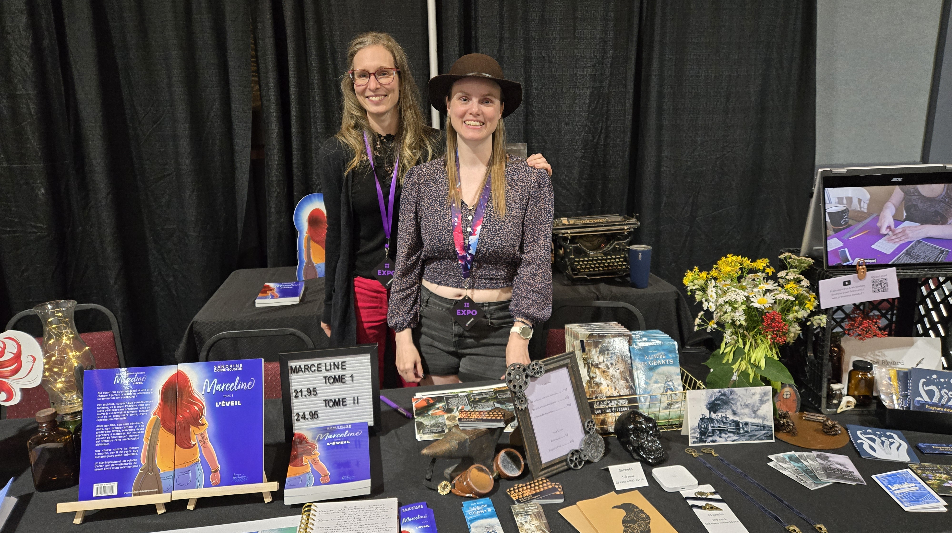 Joe Rivard et Sandrine Dionne-Gougeon se tenant derrière leur table remplie de livres et de goodies et décorée dans l'ambiance witchy steampunk