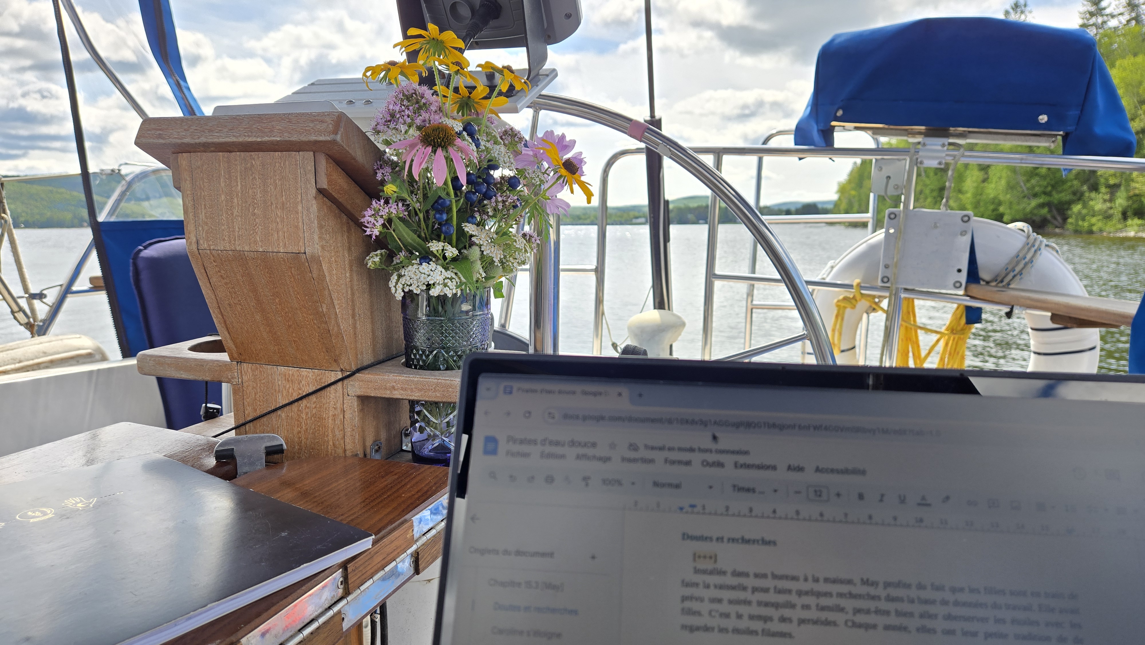 Écran d'ordinateur ouvert sur un document texte avec en arrière-plan un bouquet de fleurs et un gouvernail de voilier et la vue sur le lac Témiscouata
