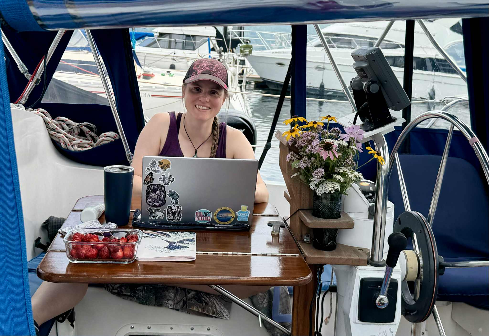 Joe assise derrière son ordinateur dans le cockpit du voilier avec en arrière-plan les bateaux de la marina