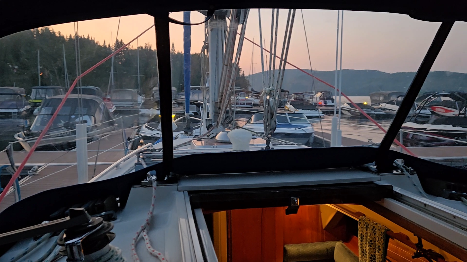 Vue de l'intérieur d'un cockpit de voilier sur les bateaux de la marina avec un ciel orangé.