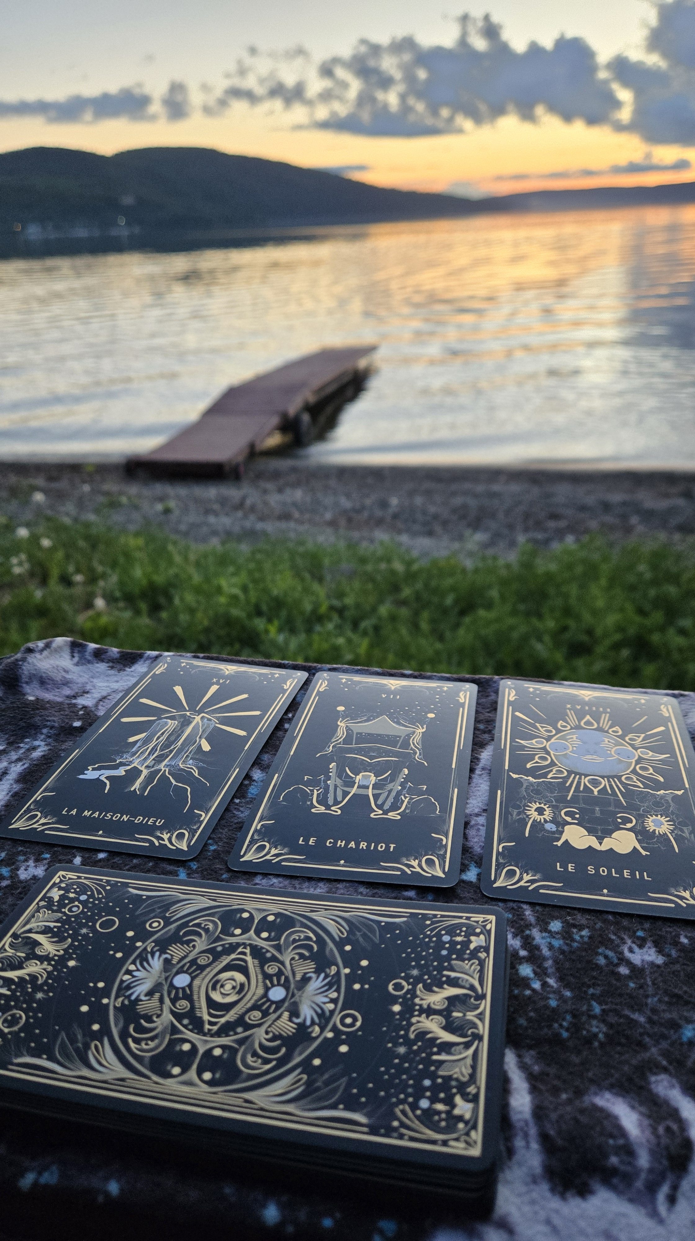 En avant-plan, trois cartes de tarot, la maison-dieu, le chariot et le soleil et en arrière-plan, un coucher de soleil sur le lac Témiscouata