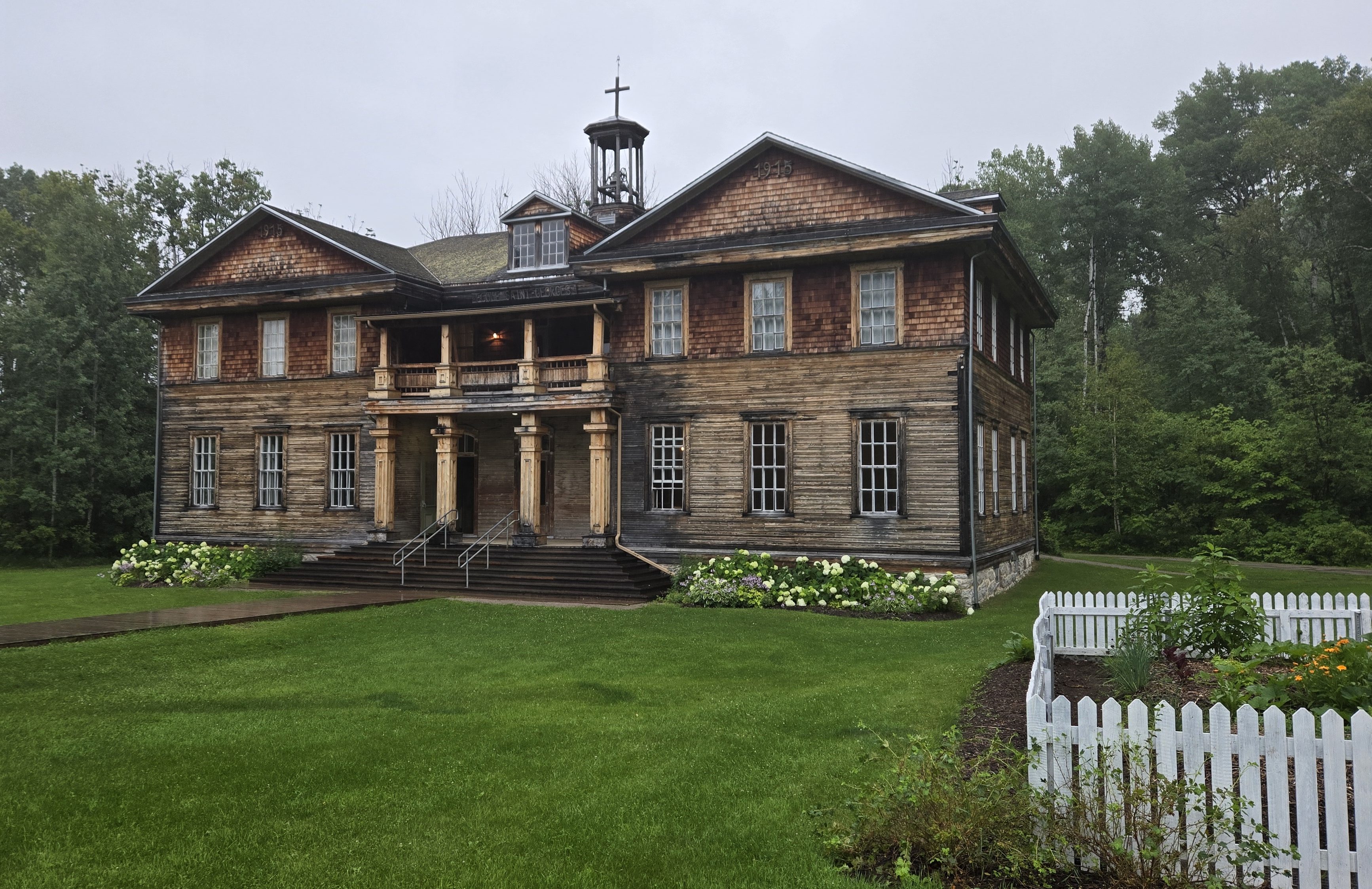 Ancienne école à deux étages en bois avec un grand porche et de grandes colonnes au centre. Les parterres sont remplis d'hydrangées et un potager à droite est entouré d'une clôture blanche.
