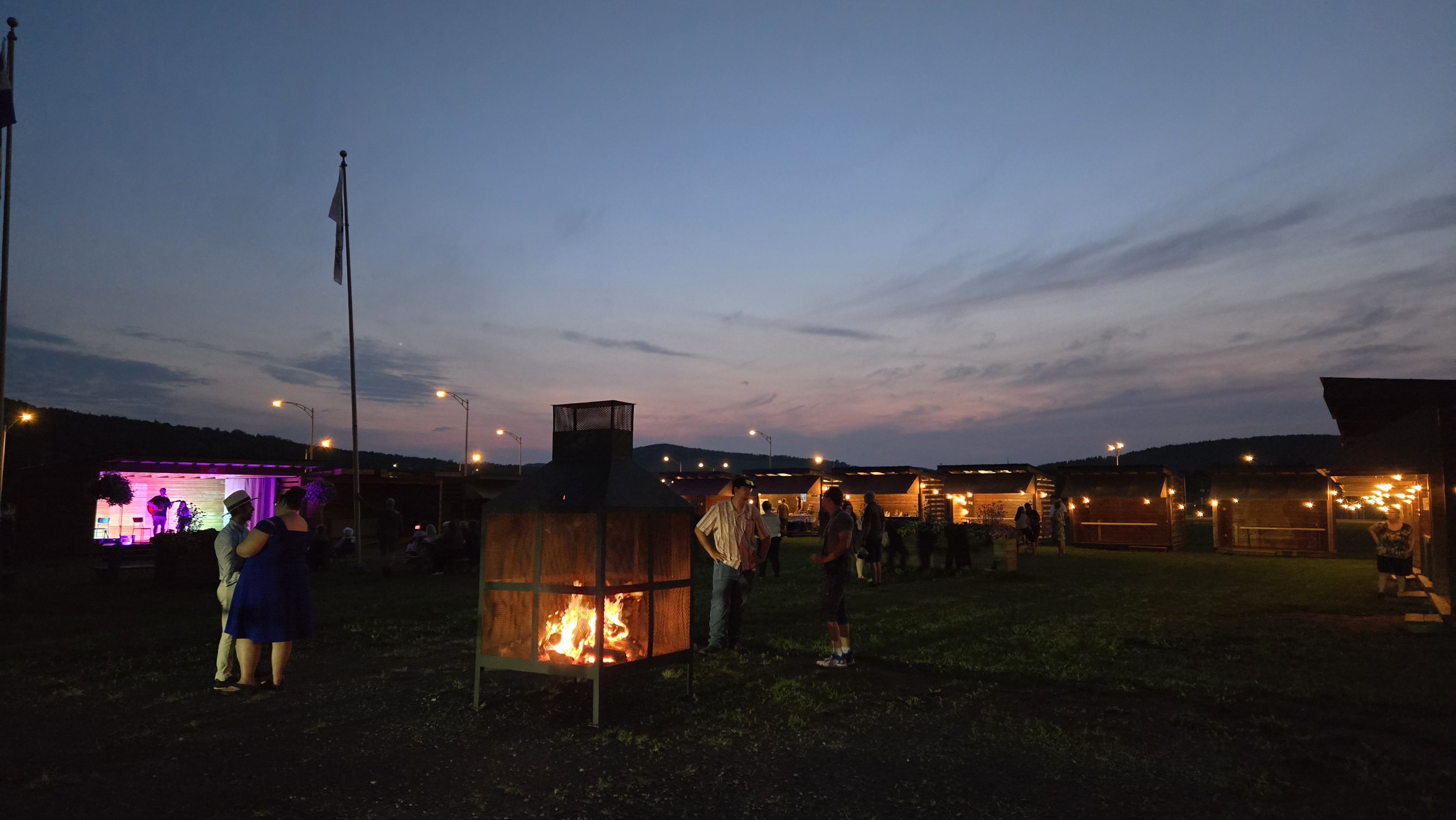 Vue sur un marché public sous une nuit naissante avec des kiosques illuminés, un feu dans un grand foyer et des musiciens sur une scène.